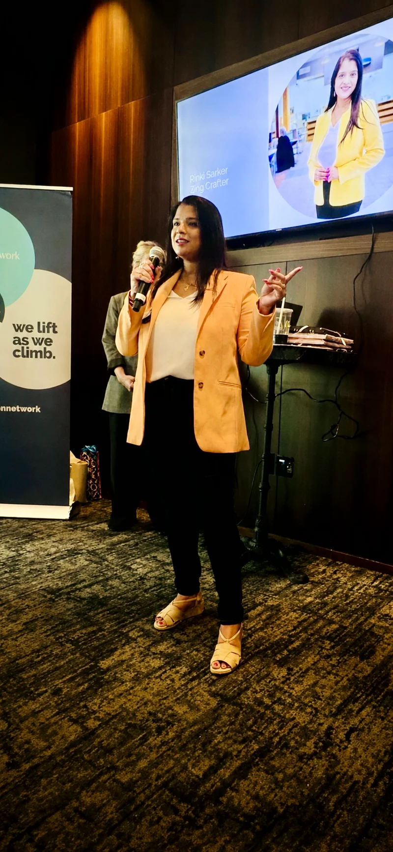 Rinki Sarker speaking into a microphone at an event while wearing a yellow jacket